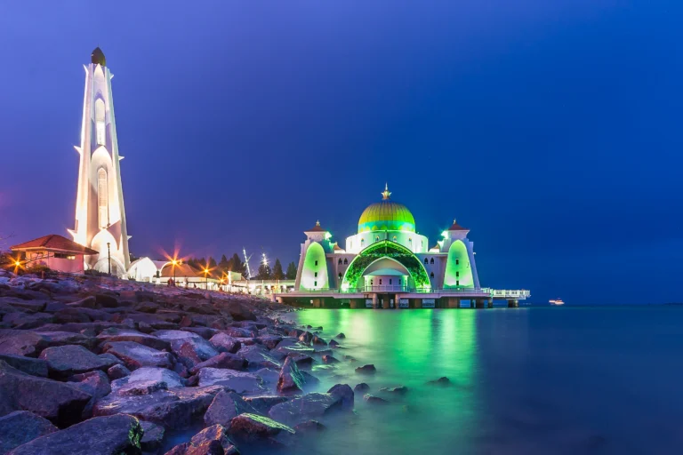 The Malacca Straits Mosque (Masjid Selat Melaka) appears to float on the sea, beautifully illuminated at night with a green dome and minaret.