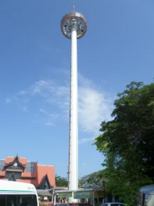 The tall white Menara Taming Sari revolving gyro tower reaching into a clear blue sky, with traditional rooftops and green trees below