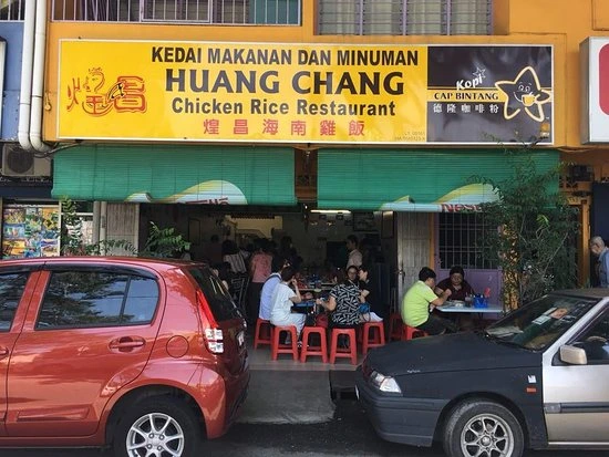 The exterior of Huang Chang Chicken Rice Restaurant with its yellow signboard, showing patrons dining at tables on the sidewalk.