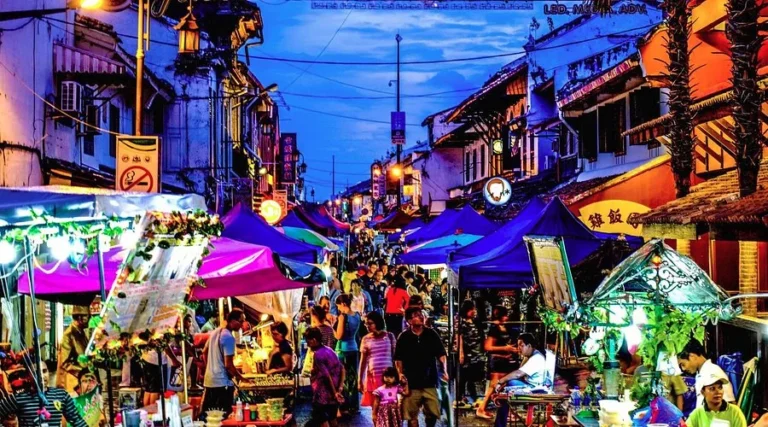 A crowded night market on Jonker Street in Malacca, with people walking between colorful food and souvenir stalls under bright lights.